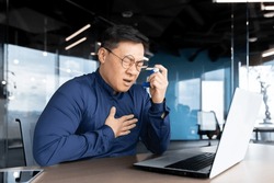 Young Asian man having asthma attack, panic attack, pneumonia at workplace. Sitting in the office at the table, holding his hands to his chest and an inhaler, he feels severe pain and is suffocating.
