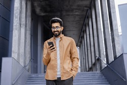 Successful young Hindu man walking outside office building, engineer software developer programmer smiling and happy using test app on phone, happy satisfied with result.