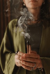A young woman holds a smoking palo santo stick in her hands. Buddhist healing practices.Clearing the space of negative energy. Aromatherapy. Selective focus,close up. Soft focus and noise effect.