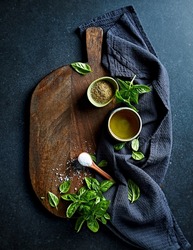 Simple arrangement of fresh basil leaves, olive oil, sea salt, ground pepper and coriander seeds on a rustic wooden chopping board. Top view