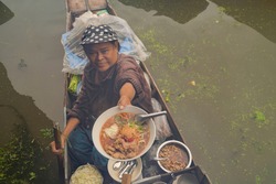 Market woman giving noodle at Damnoen Saduak Floating Market or Amphawa. Local people sell fruits, traditional food on boats in canal, Ratchaburi District, Thailand. Famous Asian tourist attraction.