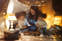 Reading and family games in children's tent. Mother and two twins daughters with books and flashlight before going to bed.