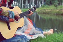 A hipster woman plays the guitar on the grass by the lake. Girl musician composes music in the evening in the country. Calm outdoor recreation on the water. Retrostyle 70s