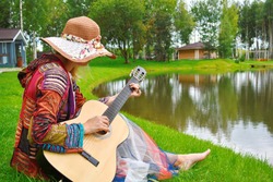 A hipster woman plays the guitar on the grass by the lake. Girl musician composes music in the evening in the country. Calm outdoor recreation on the water. Retrostyle 70s