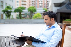 Closeup portrait, young business man deep in concentration, writing notes, sitting on wooden chair at table, isolated city background with buildings