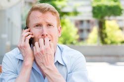 Closeup portrait, young man biting finger nails, worried about something he hears on phone, isolated outdoors background