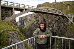 Young caucasian bearded man with curly hair posing at Mizen Head Bridge leaning on railing, County Cork, Ireland