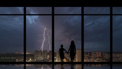 The girl and boy standing near a panoramic window against the night lightning