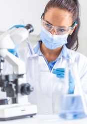 Woman in white coat, protective goggles and face mask looks at test-tube in one hand, holding a microscope slide in the other hand. Coronavirus COVID-19 concept.