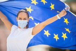 Woman in a face mask holding an European union flag behind her back with arms up.