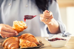 Female hands put jam onto a halved croissant in the morning.