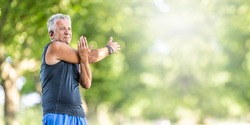 Elderly fit man stretches his arm on a summer day outdoors, wearing watches and earphones.
