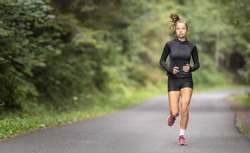 Fit young girl in black sportswear and red shoes runs on an empty asphalt road in the nature.