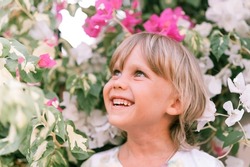 portrait of face of a candid little happy smiling five year old blonde kid boy with green eyes in pink and white flower plants in nature. children have fun the summer holidays. bright light and airy