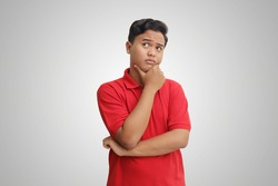 Portrait of confused Asian man in red polo shirt standing against gray background, thinking about question with hand on chin