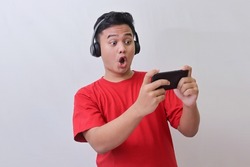 Portrait of attractive Asian man in red t-shirt using wireless headphone playing games on his mobile phone by tilting the screen. Wow face expression. Isolated image on gray background