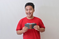Portrait of attractive Asian man in red t-shirt looking at camera, holding a wallet with rupiah banknote. Isolated image on gray background