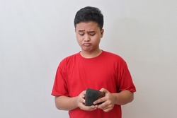 Portrait of sad Asian man in red t-shirt looking at his empty wallet. Isolated image on gray background