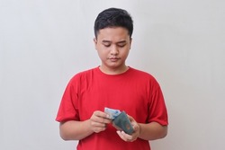Portrait of attractive Asian man in red t-shirt taking rupiah money banknote from his wallet. Isolated image on gray background