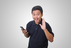 Portrait of excited Asian man in black polo shirt whispering malicious talk conversation, hand on mouth telling secret rumor while holding a mobile phone. Isolated image on white background