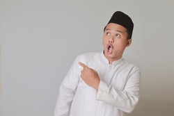 Portrait of Asian muslim man in white koko shirt with skullcap poiting to the side and showing wow face expression. Isolated image on gray background