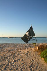 Pirate flag skull and cross bones pitched on a sandy beach at sundown with boats and sea in the background