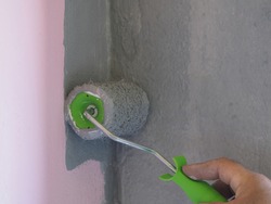 Hand holds the paint brush roller. Texture of a paint roller with light gray paint on a pink-gray wall background, closeup. Repair and painting of a concrete wall in pastel colors in a modern style
