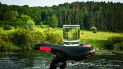 Close-up A glass of drinking water on the saddle of a sports bike, water in a transparent glass against the backdrop of a forest with a river
