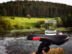 A glass of drinking water on a bicycle saddle, water in a transparent glass against the backdrop of a forest with a river