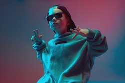 Portrait of a cool boy child in a rap image, stylishly posing in a hoodie, sunglasses and a cap on a neon background.