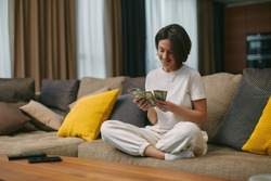 Happy young woman counting cash, enjoying money win, big profit, salary in payday sitting on sofa at home