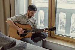 Pensive musician play acoustic guitar composing music, sitting near window at home. Focused guitarist performing melody