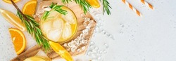 Orange juice or cocktail with rosemary and orange with ice in glass, cold summer lemonade on light gray slate, stone or concrete background. Beach concept with deep sun shadow. Top view.