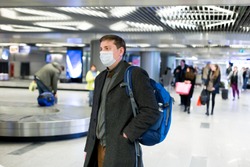 Young European man in gray coat, protective disposable medical mask in airport. Afraid of dangerous N-CoV 2019 influenza coronavirus mutated and spreading in China. Blue backpack, suitcase on wheels.