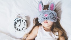 Little girl sleeping in bed under white blanket wearing grey bunny plush sleep mask with alarm clock on pillow. Early morning wake up. Putting kid to sleep.Mom's correct daily routine, rest for child.