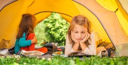 Small children, kids play,read book in orange tourist tent. Family trip, hike to nature. Backyard games, having fun. Outdoor recreation, activity. Self-assembly, setting up camping tent on lawn,grass.