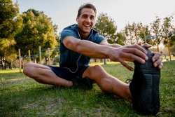 Smiling young male athlete sitting down stretching while listening to music in ear phones before exercising outdoors in park