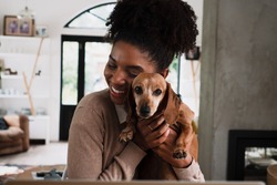 African American female smiling while snuggling cute puppy sitting at desk in modern kitchen.