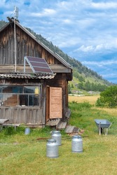 Old wooden house in mountains with solar panel on roof and milk cans on grass. The door is open. Wheelbarrow at the steps. Clouds in the sky. Vertical.