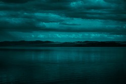   Blue green clouds over the sea. Toned seascape. Dark teal water and sky background with copy space for design.Rocky coastline on the horizon. Creepy, scary atmosphere, mood.                         