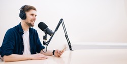 Man recording podcast in white studio. hand holding mobile phone in audio home studio. Male with headphones doing online meeting. Remote radio host broadcasting from home with professional microphone.