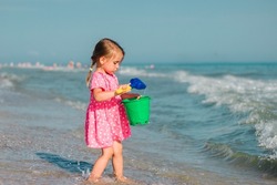 A little girl plays with a shoulder blade and a bucket on the beach in a pink heart dress. 