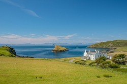 View of a white house among green fields and hills with small rocky island on the ocean behind it, Isle of Skye, Scotland, United Kingdom

