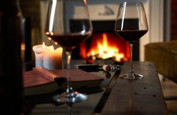 Two glasses of red wine with book and candle on table at home, fireplace in the background. Warm, dark colors.