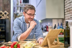 Handsome older man wearing eyeglasses, cooking at home in the kitchen, cutting vegetables on board, following recipe in online cookbook app, using tablet. 