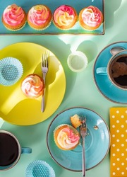 Beautiful colourful and bright table setting with food coloured cup cakes and cyan blue coffee cups, yellow dotted napkin, and green plate arranged in a geometrical style