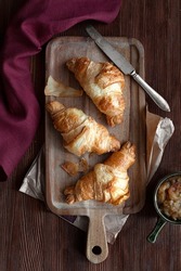 Beautiful food photography with croissants on a wooden kitchen board, knife, purple linen fabric towel and jam behind on a dark wooden table