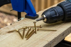 cross head countersunk brass single thread wood screws on a wooden board plank with an electric drill screwdriver and clamp outside