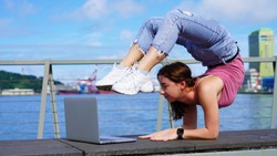 beautiful flexible girl stands in a bridge in front of a laptop on a viewing platform near the port. Lifestyle girl working on a journey. Backbend pose.