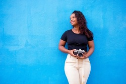 Young happy black woman taking pictures with her camera in front of a blue wall.